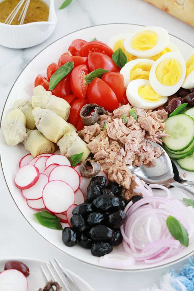 Dans un plat de service : ingrédients de la salade niçoise classique : tomates, oeufs durs, thon, anchois, olives, artichauts, oignon, radis, concombre, huiile d'olive, vinaigre, sel et poivre.