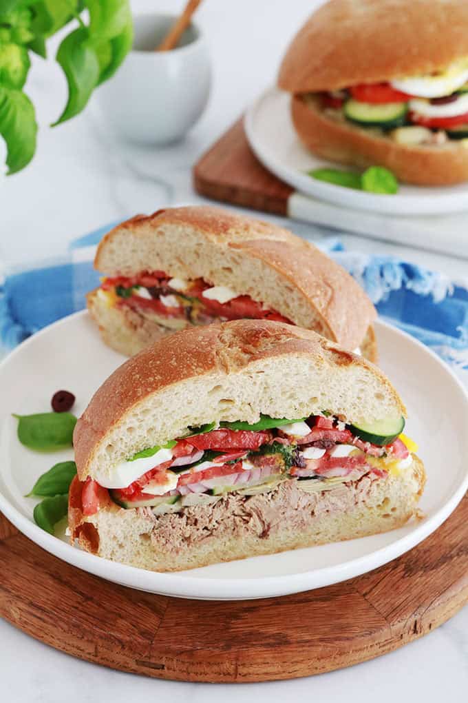 Dans deux assiettes, Sandwichs Pan bagnat, pains ronds garnis de salade niçoise (thon, tomates, oignon, concombre, artichaut, oeufs durs, anchois, olives et basilic.