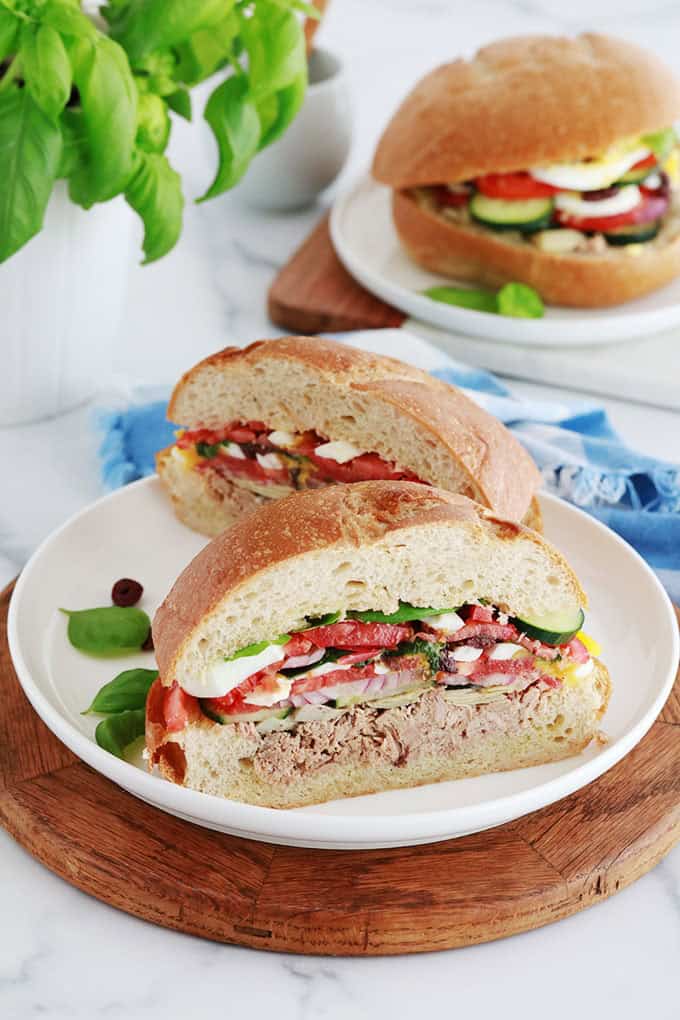 Dans deux assiettes, Sandwichs Pan bagnat, pains ronds garnis de salade niçoise (thon, tomates, oignon, concombre, artichaut, oeufs durs, anchois, olives et feuilles de basilic).