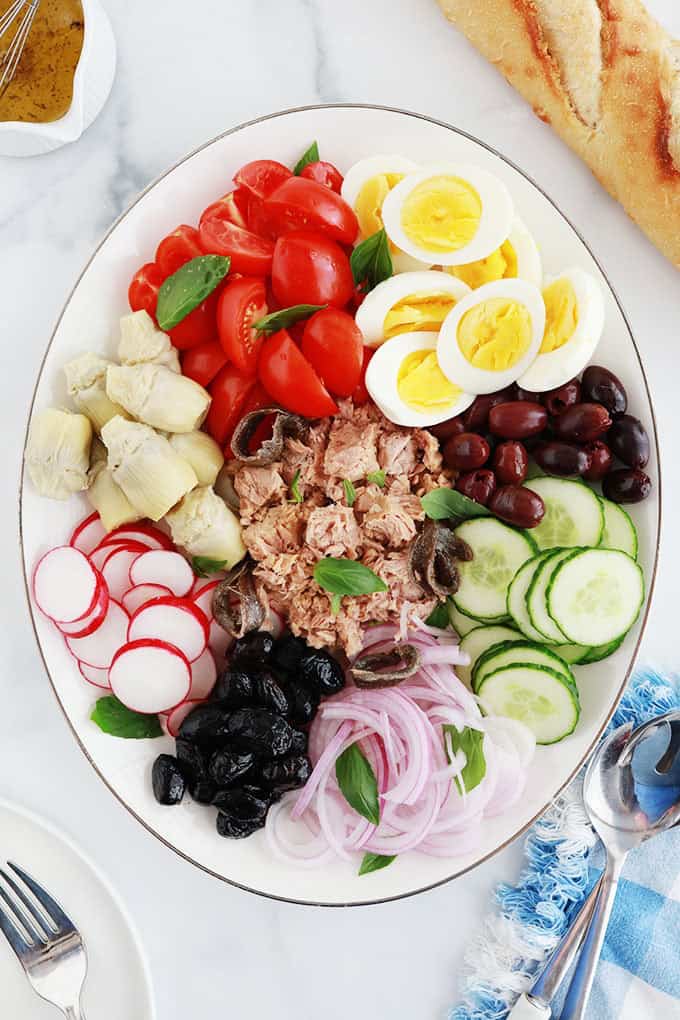 Dans un plat de service : ingrédients de la salade niçoise classique : tomates, oeufs durs, thon, anchois, olives, artichauts, oignon, radis, concombre. Dans un petit bol, une vinaigrette simple. Pain baguette. 